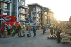 A group of construction workers sit by piles of wood and cranes on a new apartment complex construction, while the sun glares over them.